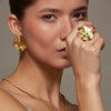 A close-up portrait of a model wearing a coordinated gold jewelry set with textured organic-shaped pieces, including a ring, earrings, necklace, and bracelets, accented with Colombian green stones.