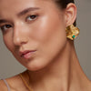 Close-up of a woman wearing gold earrings with one colombian emerald, against a neutral gray background.